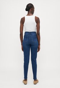 Høytlivede blå denimjeans med slim fit, med baklommer og en lett stretch, parret med en hvit tanktop og lyse sko.