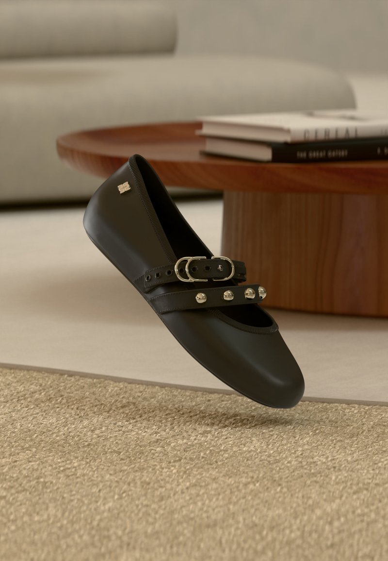 Chaussure plate en cuir noir avec double lanières à boucles et clous métalliques, suspendue en l'air au-dessus d'un tapis beige texturé et d'un fond de table en bois.