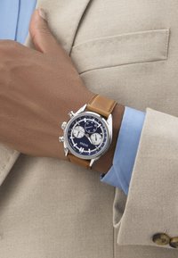 Polso con orologio cronografo Guess a cinturino in pelle marrone sopra la manica di una camicia blu e un blazer beige, mano appoggiata sul petto.