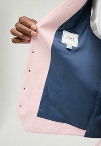 Interno di un blazer rosa chiaro con una fodera blu scuro liscia. Presenta un’etichetta con la scritta "NEXT TAILORING" e diversi piccoli accenti di filo nero.