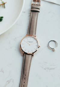 Orologio rotondo con un quadrante bianco, dettagli dorati e un cinturino in pelle grigia chiaro. Include un anello argento con una piccola perla accanto.