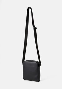 Bolso cruzado de cuero negro con una textura suave, forma rectangular, correa de nylon ajustable y cierre con cremallera.