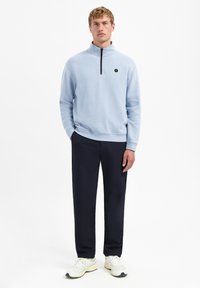 Lichtblauwe fleece trui met een kwart rits, voorzien van een zwarte rits en een klein logo op de borst. Combinatie met donkere broek en witte sneakers.
