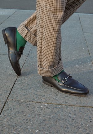 Czarne skórzane mokasyny z klamrą, zestawione z zielonymi skarpetami i brązowymi spodniami w kratę, ustawione na kamiennej nawierzchni.