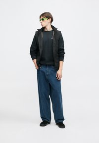 Melna pūšamā veste pār melnu sporta džemperi, kopā ar vaļīgām zilām džinsa biksēm. Zaļas saulesbrilles piešķir krāsas akcentu. Melni apavi noslēdz izskatu.