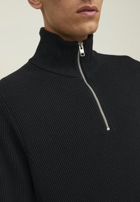Maglione nero a coste con collo alto e zip argentata, caratterizzato da un design aderente e un tessuto testurizzato. Vista ravvicinata che mette a fuoco il collo e il dettaglio della zip.