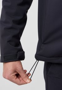 Veste softshell noire avec cordon de serrage ajustable à l'ourlet, dotée d'une fermeture à velcro aux poignets et d'un matériau à texture lisse.