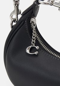 Mustakäsineinen nahkalaukku, jossa on kaareva muoto, sisältäen hopeanvärisen vetoketjun ja ketjuyksityiskohtia, joita korostaa metallinen riipus. Silkinomainen pinta.