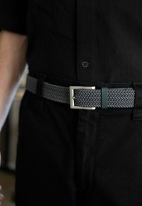 Geflochtener grauer Gürtel mit einer silbernen Metallschnalle. Strukturiertes Gewebedesign, gesichert mit einem grünen Akzent in der Nähe der Schnalle. Über schwarzer Kleidung getragen.