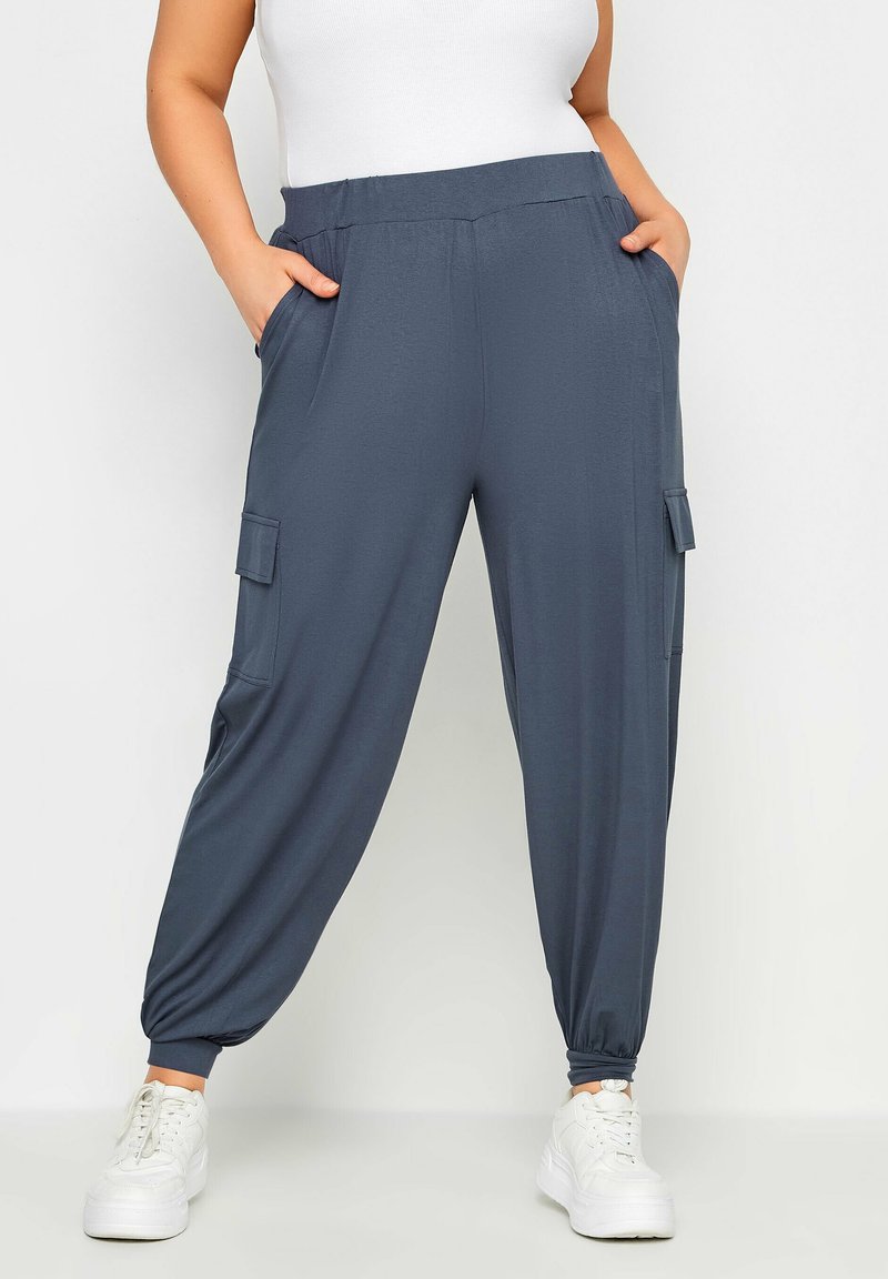 Femme portant un pantalon cargo jogger bleu avec des poignets élastiques et des baskets blanches, les mains dans les poches, devant un fond uni.