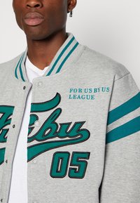 Gray sweatshirt jacket with teal embroidered logo and stripes, featuring snap buttons and “FOR US BY US LEAGUE” text on the chest.