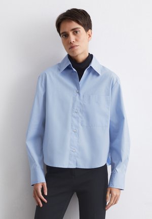 Camisa de color azul claro, corta, con botones y cuello puntiagudo, un bolsillo en el pecho y mangas largas. Presenta una tela ligera y un ajuste relajado.