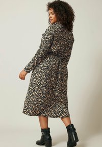 Kleid mit wellenförmigem Muster in Beige und Schwarz, langärmelig mit tailliertem Bund. Getragen mit schwarzen Knöchelstiefeln mit Schnallendetail.