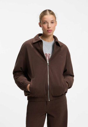Jonge vrouw die een bruine jas met rits draagt, met handen in de zakken en bijpassende broek, kijkt recht vooruit met een neutrale uitdrukking.