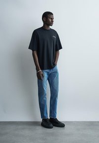Homme debout portant un t-shirt noir oversize, un jean bleu clair, des baskets noires et un bracelet argenté devant un mur uni gris clair.