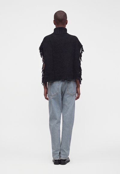 Personne aux cheveux courts portant un poncho noir à franges et un jean bleu clair, debout de dos sur un fond uni.