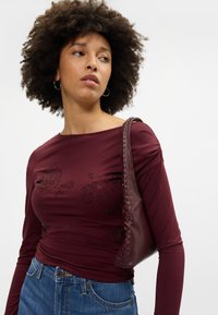 Garšīgas piedurknes bordo krāsas blūze ar melnu ziedu rakstu un rotājumiem; saskaņota ar tumši brūnu ādas plecu somu ar austiem elementiem.