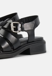 Μαύρο δερμάτινο σανδάλι με πολλαπλούς ιμάντες, ασημί πόρπη κλεισίματος, επένδυση πέλματος με μαξιλαράκι και χαμηλό στρωματοποιημένο τακούνι σε λευκό φόντο.