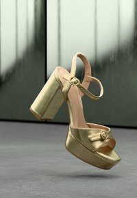 Χρυσό μεταλλικό δίπατο πέδιλο με υψηλά τακούνια, λουριά στον αστράγαλο και τα δάχτυλα με χρυσά κουμπώματα σε ανακλαστικό φόντο.