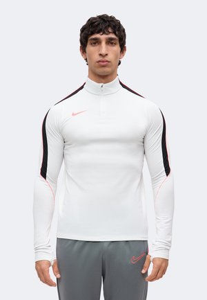 Ung mand iført en hvid langærmet sportstop med sorte og røde striber og grå bukser med et rødt Nike-logo.