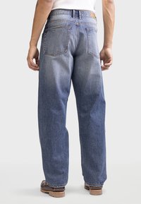 Jeans de denim azul con un ajuste relajado, dos bolsillos traseros y una textura desgastada. Piernas ligeramente entalladas con una cintura clásica y detalles en marrón.