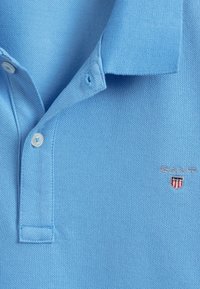 Lyseblå polo-shirt lavet af struktureret bomuld, med en treknaps lukning og et lille broderet GANT-logo med skjolddetalje.