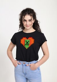 Černé tričko s krátkým rukávem s pixelovanou zelenou postavou uvnitř oranžového srdce, uvolněný střih a měkký materiál.