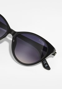 Lunettes de soleil cat-eye noires avec des verres sombres dégradés, présentant une monture brillante et le nom de la marque "GUESS" en argent sur le verre gauche.