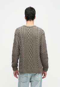 Mann in braunem, grob gestricktem Pullover und hellblauen Jeans, mit dem Rücken zu einer schlichten weißen Hintergrund.