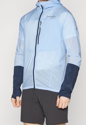 Jasnoniebieska kurtka z kapturem, z ciemnogranatowymi dolnymi rękawami i czarnym zamkiem błyskawicznym, noszona z czarnymi szortami przez osobę stojącą na białym tle.