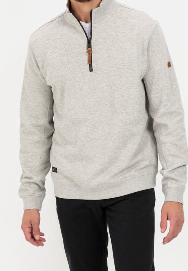 Homme portant un pull gris clair à fermeture éclair quart de zip avec manches longues et pantalon noir, debout devant un fond uni.