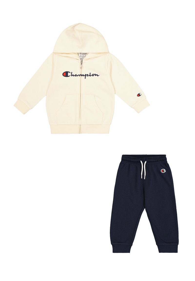 Felpa crema con zip e logo "Champion" blu, abbinata a pantaloni della tuta blu con vita elastica e toppa con logo sulla gamba.