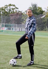Sportovní sako s geometrickými vzory v modré a bílé, černé kalhoty se světle šedými akcenty a černé fotbalové boty. Fotbalový míč na trávě.