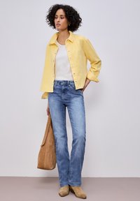 Femme portant une veste jaune boutonnée, un haut blanc, un jean bleu, des bottes couleur miel, tenant un grand sac en daim couleur miel, debout contre un mur clair uni.