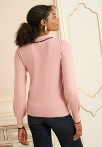 Rosa Strickpullover mit langen, puffartigen Ärmeln und einem gewellten Ausschnitt. Besitzt einen dunklen Besatz an den Bündchen und dem Saum. Wird mit marineblauen Hosen getragen.