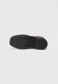 Brun lädersandal med en svart gummisula. Har en texturerad yta och ett litet spänne för att fästa foten.