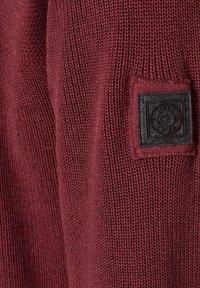 Burgunderfarbener Strickpullover mit strukturiertem Stoff, tailliertem Schnitt und einem quadratischen schwarzen Patch mit geprägten Details an der Seite.