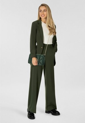 Donna con capelli lunghi biondi che indossa un completo verde oliva e una maglietta bianca, tiene in mano una borsa trapuntata verde con tracolla a catena, in piedi su sfondo bianco.