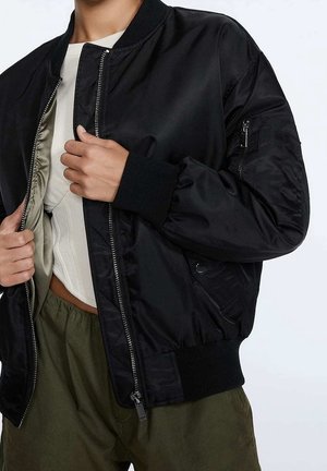 Personne portant une veste bomber noire non zippée sur un haut blanc et un pantalon vert, ajustant la veste avec les deux mains.
