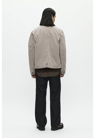 Beige denimjack met schoudernaden, rugventilatie, gedragen over een donkergroene geruite shirt, gecombineerd met zwarte losse broeken en sandalen.