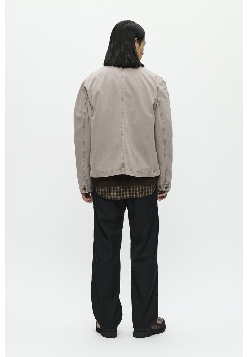 Beige denimjakke med skuldrebrede sømme, rygventilation, båret over en mørkegrøn ternet skjorte, parret med sorte løse bukser og sandaler.