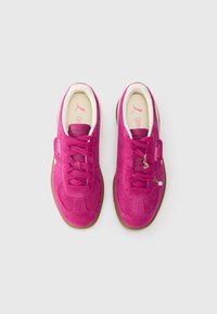 Un paio di sneakers Puma in suede rosa brillante con suole in gomma, fodera interna bianca e piccoli ciondoli a forma di cuore dorati sui lacci della scarpa destra.