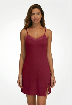 Vrouw die een mouwloze bordeauxrode jurk draagt met kanten rand en een kleine zijscheur, staand tegen een effen witte achtergrond.