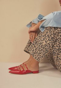 Mules en cuir rouge avec des pointes et trois lanières. Associés à un pantalon large en imprimé léopard, mettant en valeur le contraste de texture et de couleur.
