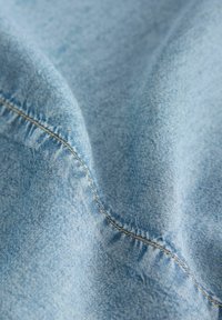 Ljusblå denimtyg med en slät yta, som har synliga sömmar längs kanterna och lätt färgvariation.