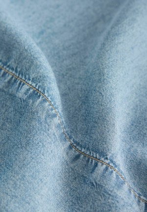 Helles, blaues Denim-Gewebe mit glatter Textur, das sichtbare Nähte entlang der Kanten und leichte Farbvariationen aufweist.