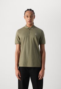 Polo shirt de culoare verde măsliniu, cu mânecă scurtă, din material cu textură ribbed. Are guler cu trei butoane și un mic logo brodat pe pieptul stâng. Pantaloni negri purtați.