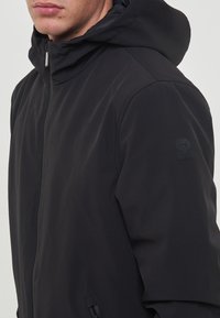 Giacca nera con cappuccio realizzata in un tessuto liscio e opaco, con chiusura a zip e un accento logo sulla manica. Progettata con una silhouette moderna ed elegante.