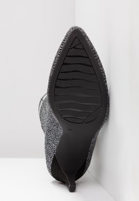 Chaussure à talon haut noire et argentée avec un dessus en tissu texturé, bout pointu et une semelle en caoutchouc présentant des rainures horizontales. Taille marquée comme 37.