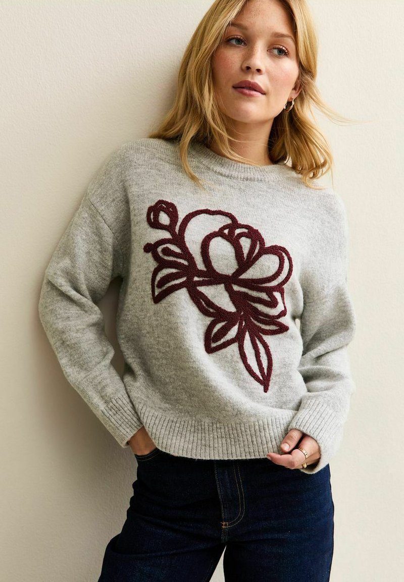 Maglione grigio lavorato a maglia con orlo a costine, caratterizzato da un grande motivo floreale ricamato in bordeaux sul davanti. Abbinato a pantaloni jeans scuri.